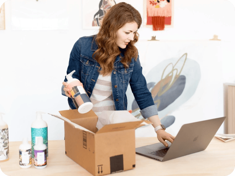 Woman holding a bottle over an open cardboard box while using her laptop. 