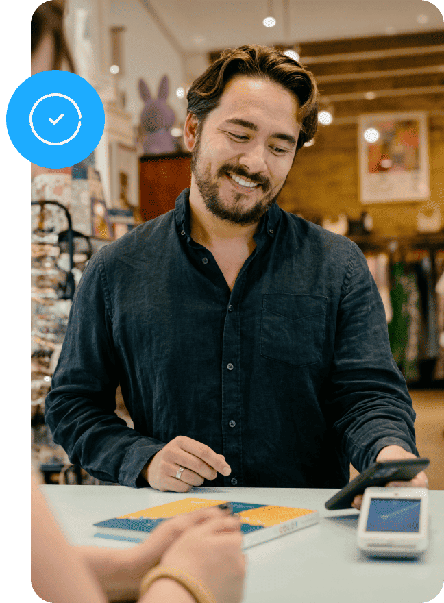 An offscreen customer taps their card to pay for a purchase on the customer-facing side of a Genius countertop POS. A small business employee stands on the other side of the counter tapping the employee-facing screen of the POS.