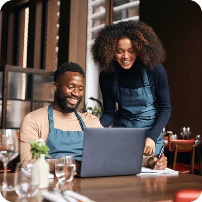 Two small business employees in aprons smiling while looking at an open laptop in a restaurant