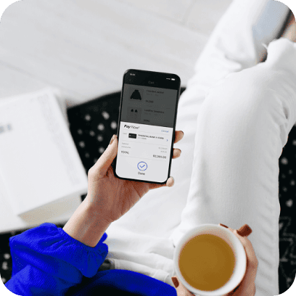 Overhead shot of a person completing an online transaction on their phone using Apple Pay. They are sitting on a couch and holding a cup of coffee