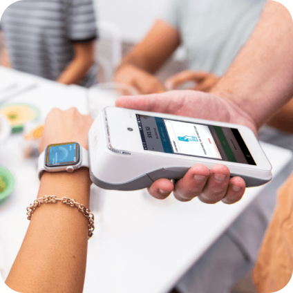 A person pays with a smartwatch by holding their wrist near a card reader, while others sit at a table with food in the background.
