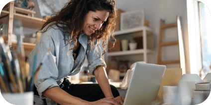 A woman leans over a table, smiling while using a laptop in a workspace filled with art supplies and ceramics.