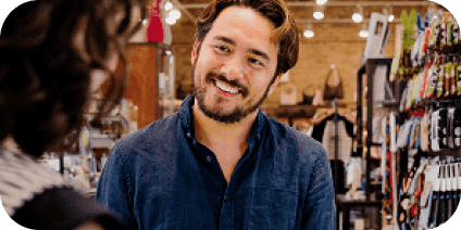 A man with a beard and blue shirt smiles and talks to a customer inside a retail store. Shelves with various merchandise are visible in the background.