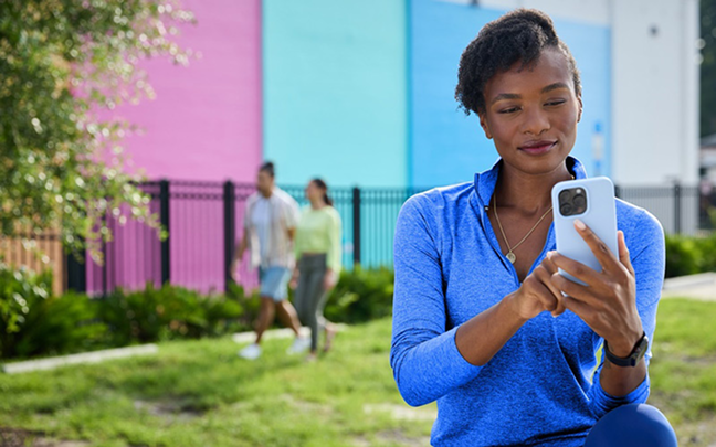 A woman in a blue top uses her smartphone outdoors, with a colorful wall and two people walking in the background.