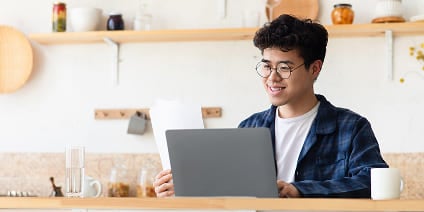 Young man with glasses reviewing documents while using a laptop in a kitchen.