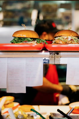 Two gourmet burgers on red trays at a restaurant pass with hanging order tickets.