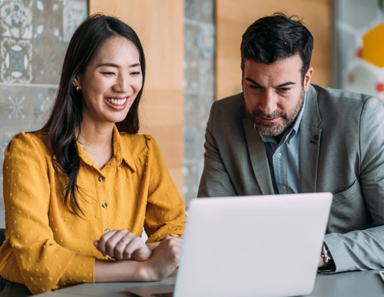 A woman and man collaborating at a table while reviewing a laptop.