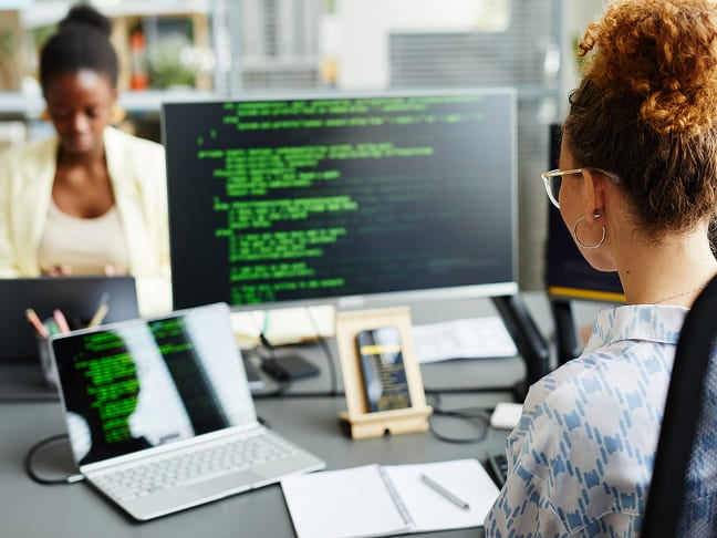 A woman reviewing lines of code on dual screens at her desk setup