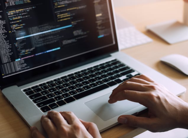 Close-up of hands operating a laptop displaying programming code on the screen