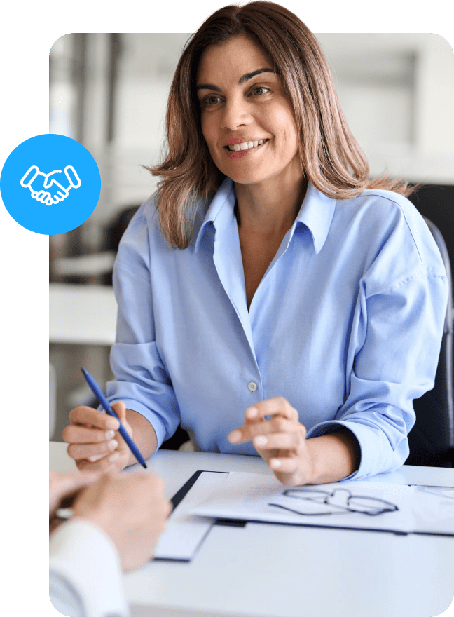 A smiling woman in a light blue shirt sits at a desk holding a pen, with a blue circle icon featuring a white handshake symbol in the top left corner.