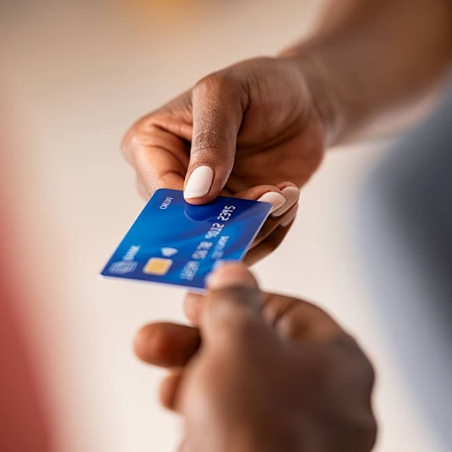 A close-up view of two hands exchanging a blue credit card with a gold chip, held between fingers against a soft, blurred, light-colored background.