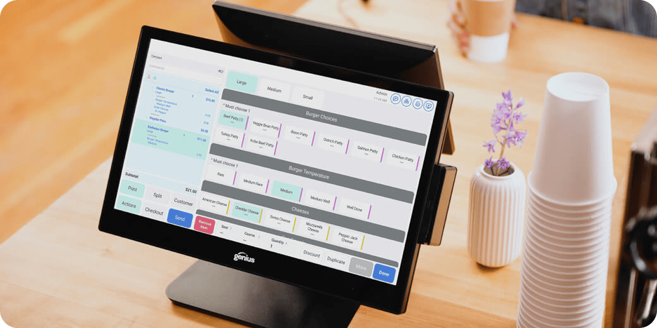 A point-of-sale touchscreen display showing a burger ordering menu, placed on a wooden counter next to a stack of white cups and a small flower vase.