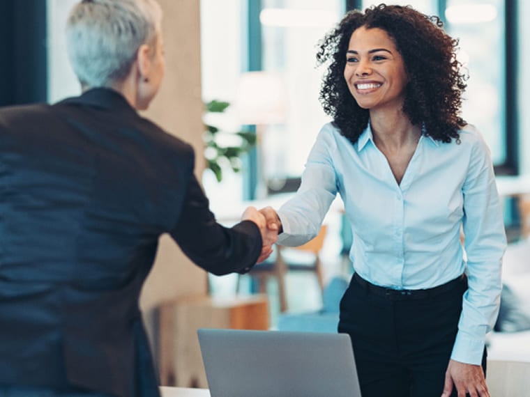 Two business professionals shaking hands across a desk near a laptop.