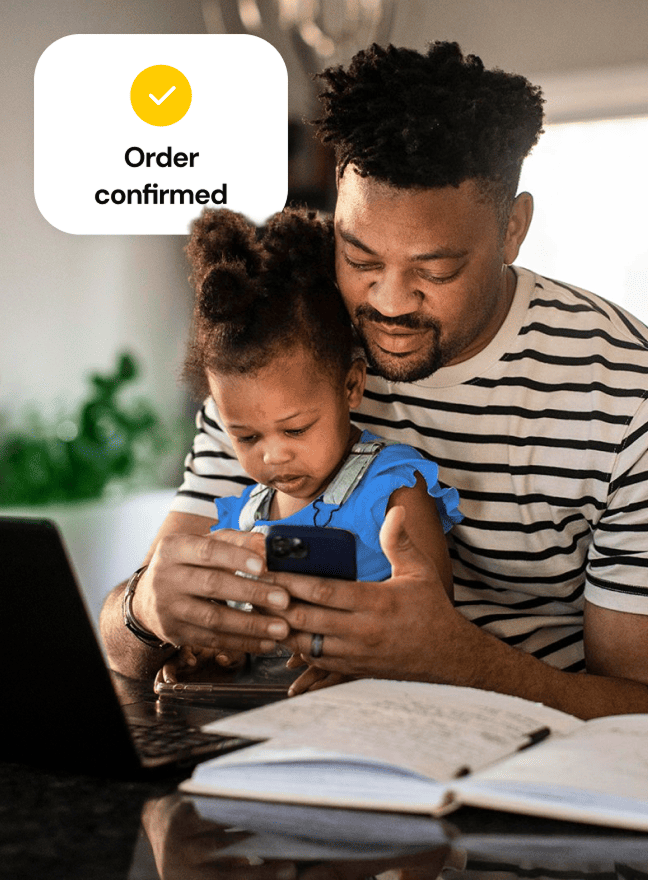 A man sitting at his home in front of an open laptop holds his daughter in his lap as he looks at a phone, while a small graphic overlaying the image shows an order confirmed graphic.