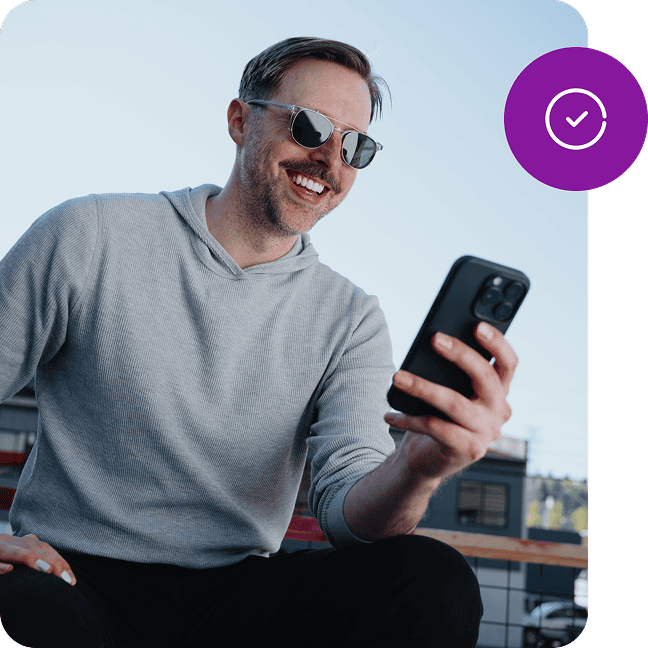 A smiling man in sunglasses looks down at a phone in his hand and a small, round, graphic icon showing a checkmark overlays the image.