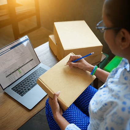 Person writing on a cardboard box next to a laptop displaying order data.