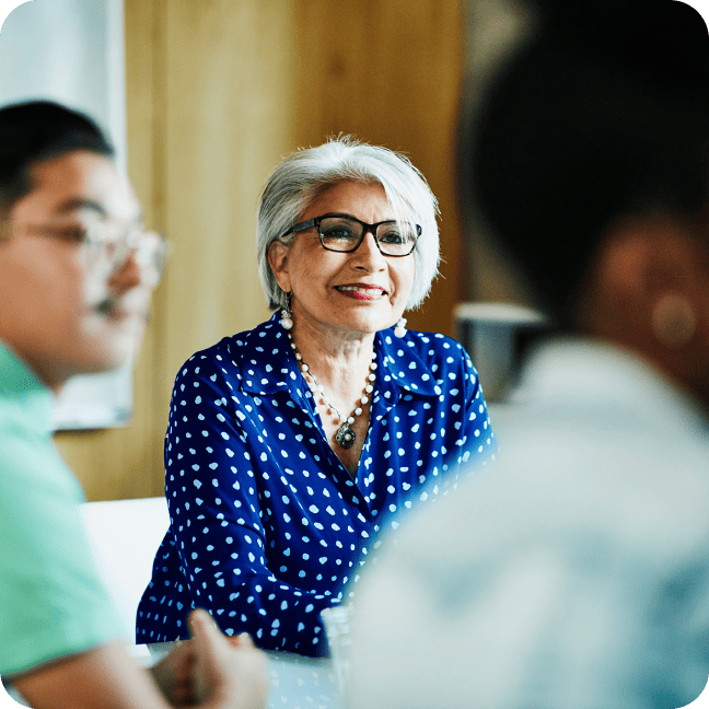 An older woman with grey hair and glasses listening attentively in an office setting.