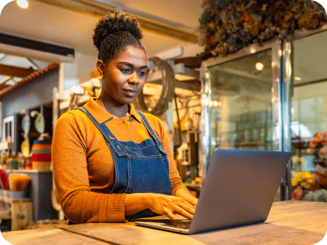 Woman working on a laptop computer at a shop counter in a cozy store setting.