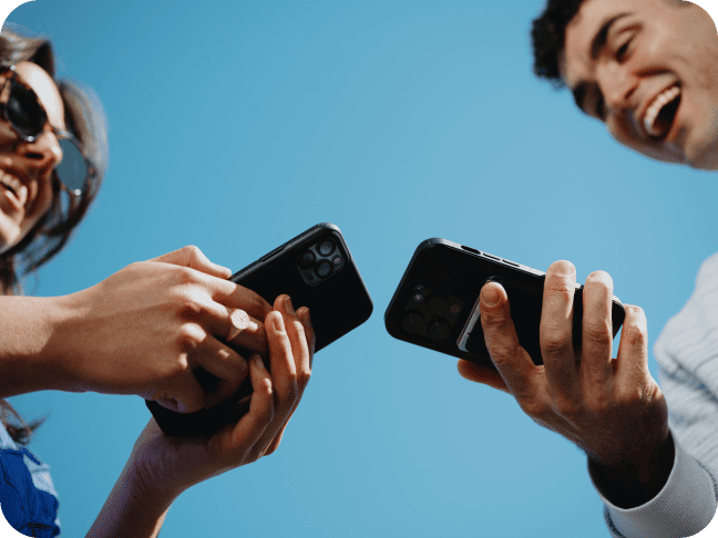 Low-angle shot of two smiling people holding smartphones together against a blue sky.