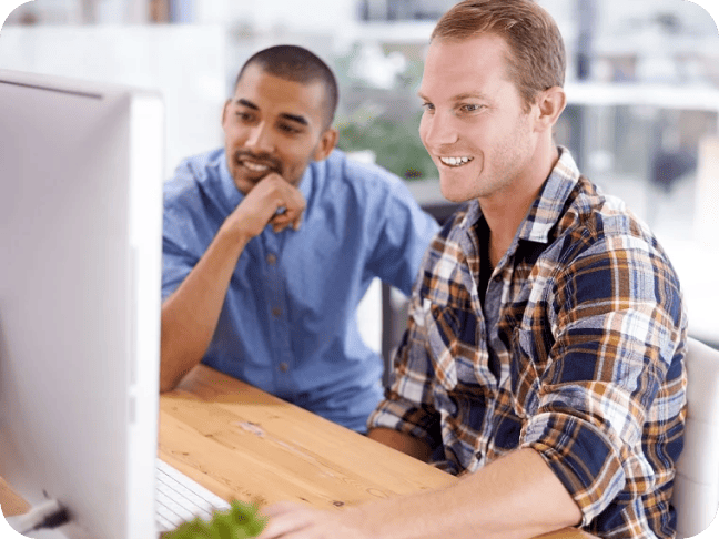 Two men sit at a desk looking at a computer monitor, one smiling and using the mouse while the other observes with interest.