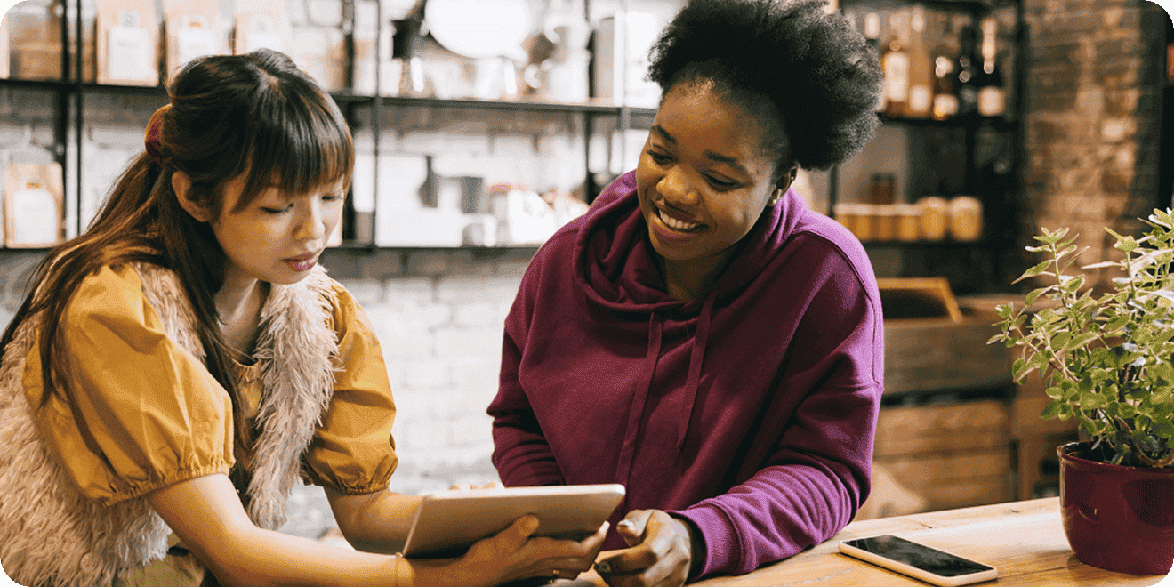 Two women sit at a wooden table in a cozy cafe, looking at a tablet together. One woman is showing something on the tablet while the other smiles. A smartphone and plant are on the table.