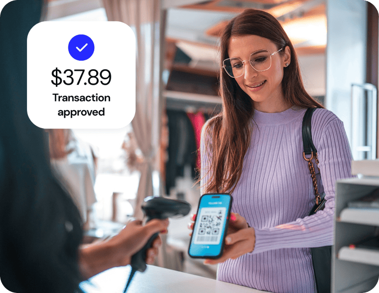 A woman in a store holds her cell phone, displaying a QR code. The cashier scans the QR code to complete the transaction.