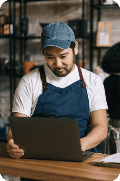 Man in a cap and blue apron typing on a laptop computer in a café setting.