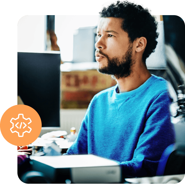 A man in a blue sweater focused at his computer desk with a coding icon graphic.