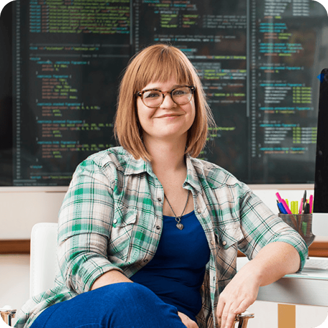 A smiling woman with glasses in a plaid shirt sitting in front of screens filled with code.