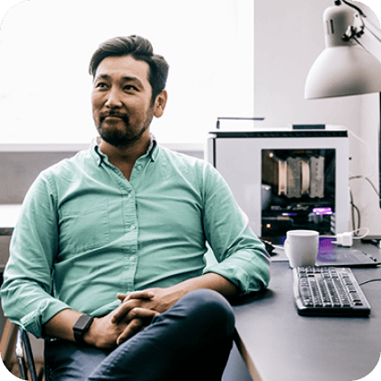 A man in a green shirt sitting thoughtfully at his office desk next to a desktop PC.