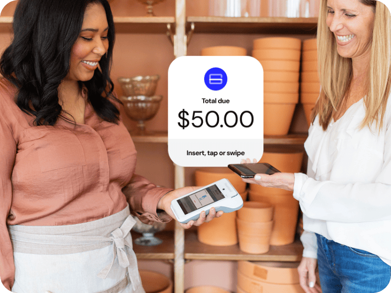 Two women smile in a flower shop as one makes a payment via smartphone to a handheld device. 