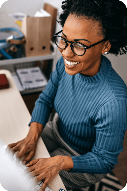 A smiling woman wearing a blue sweater and glasses works at her desk.