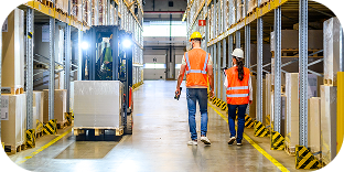 Two people wearing orange safety vests and hard hats walk inside a warehouse.