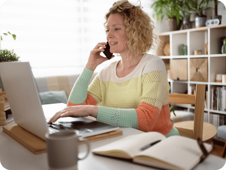 A woman in a colorful sweater sits at a desk using a laptop while talking on a phone.