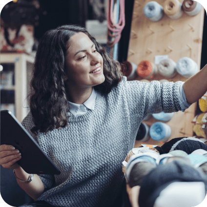 A smiling woman holding a tablet while reaching for yarn in a craft shop.