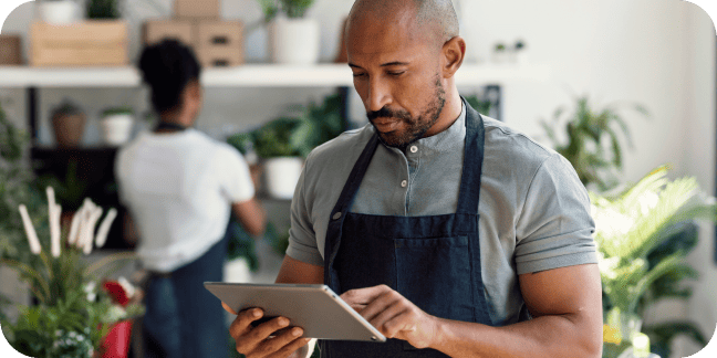 Plant shop worker in an apron using a tablet with a colleague in the background.