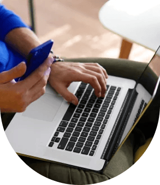 Person holding a blue smartphone in one hand while typing on a laptop with the other.