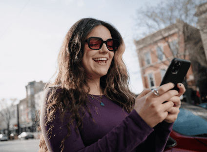 A smiling woman wearing sunglasses using her smartphone on a city street.