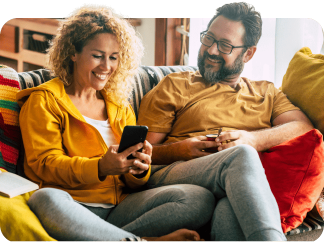 Happy couple sitting on a sofa at home looking at a mobile phone together.