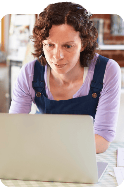 Woman wearing a purple shirt and apron leaning over to view her laptop.