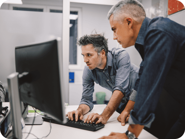 Two men in an office work together at a computer; one types while the other leans over to view the screen.