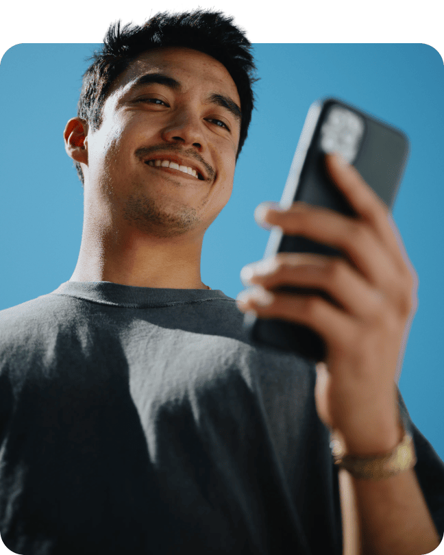 A young man smiling warmly while using his smartphone outdoors under a clear blue sky.