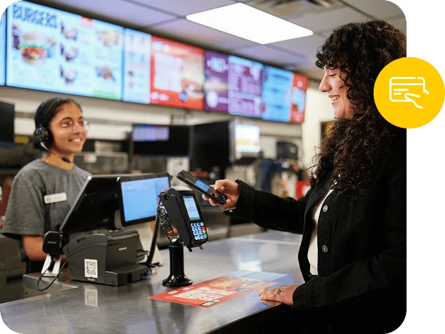 A digital menu board lights up the background behind a fast food worker, accepting a point of sale payment.