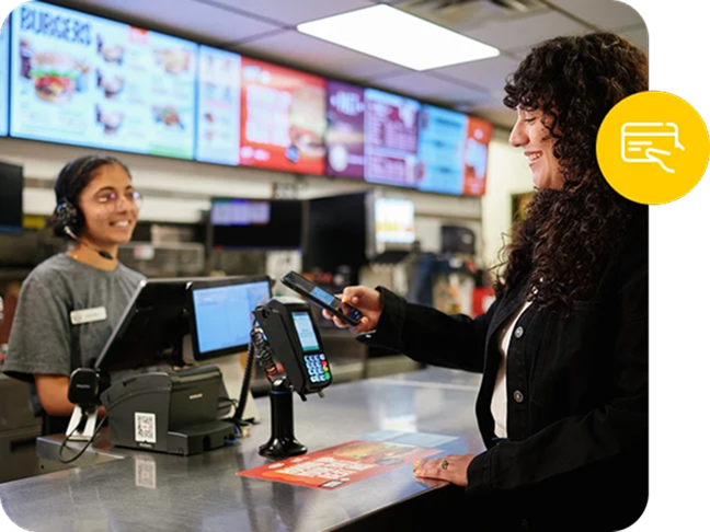 A digital menu board lights up the background behind a fast food worker, accepting a point of sale payment.