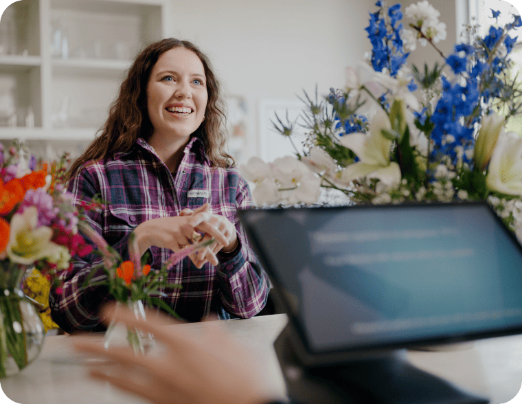 A woman smiling behind a store counter with vibrant floral displays and a digital POS screen.