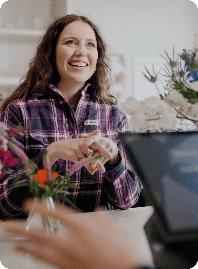 Smiling woman sitting at a retail desk with a blurry touchscreen tablet in the foreground.