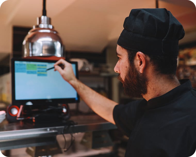A person standing in the back-of-house wearing a chef’s coat and hat reviewing ticket items via screen connector.