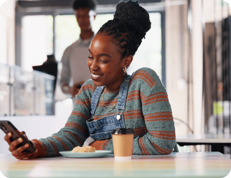 Customer smiling while looking at a smartphone at a café table with a coffee cup and snack.