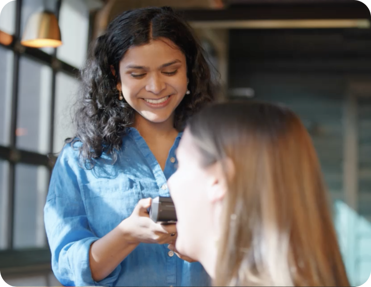 A person in a blue shirt smiling and holding a payment terminal toward another seated person in a well-lit indoor setting