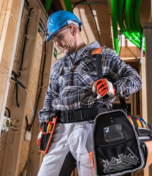 A contractor wearing a hardhat and toolbelt holds a large toolbag and observes a project in the progress. 